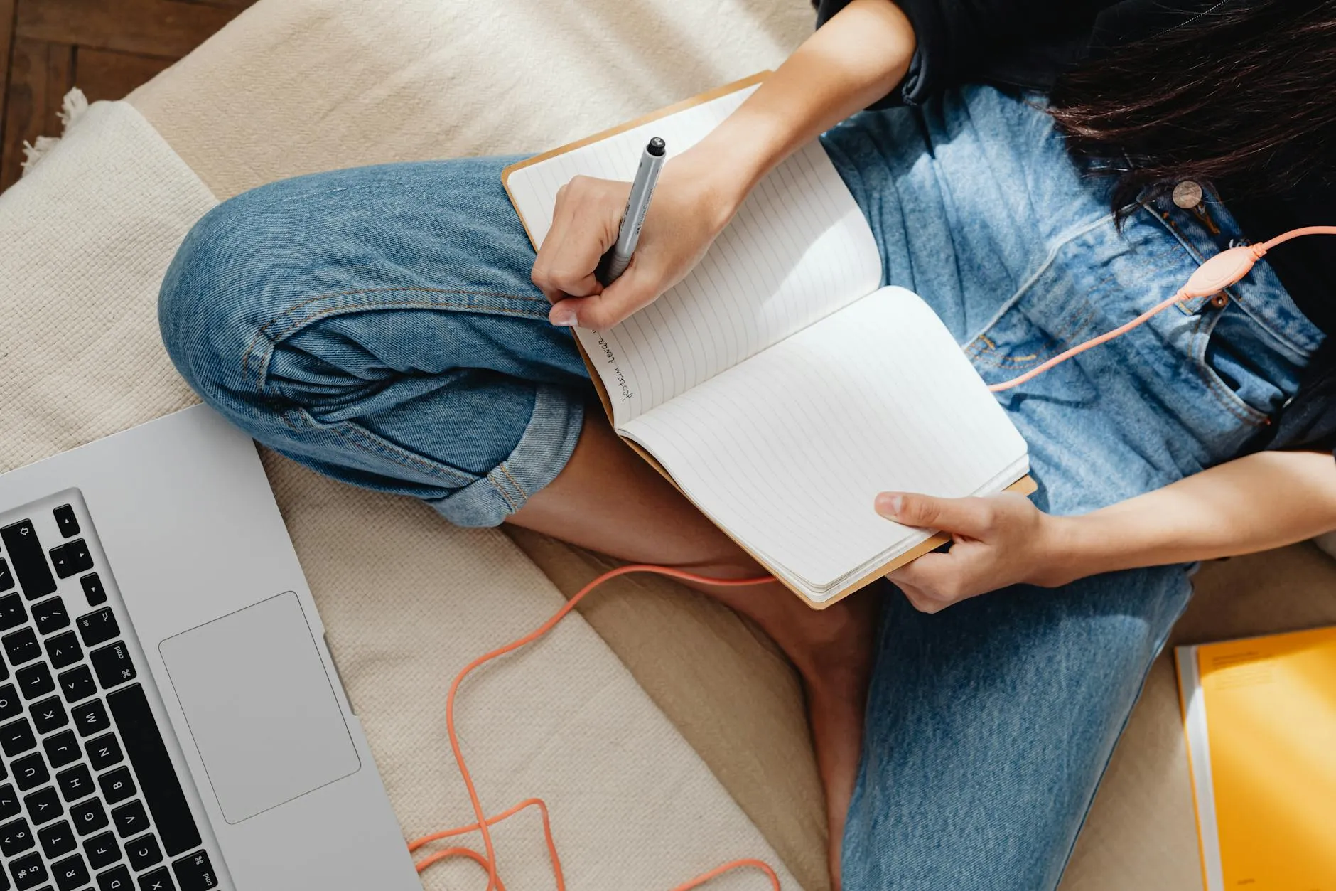 Pessoa escrevendo projeto acadêmico em caderno com laptop aberto sobre mesa de madeira