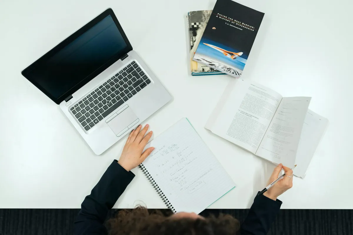Pessoa digitando em laptop com documentos acadêmicos ao lado, em ambiente de estudo.