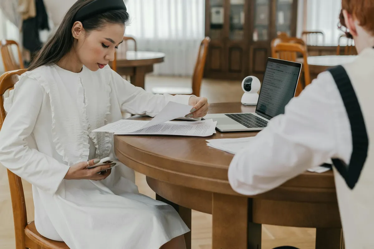 Pessoa lendo documentos acadêmicos em mesa de trabalho com computador ao fundo