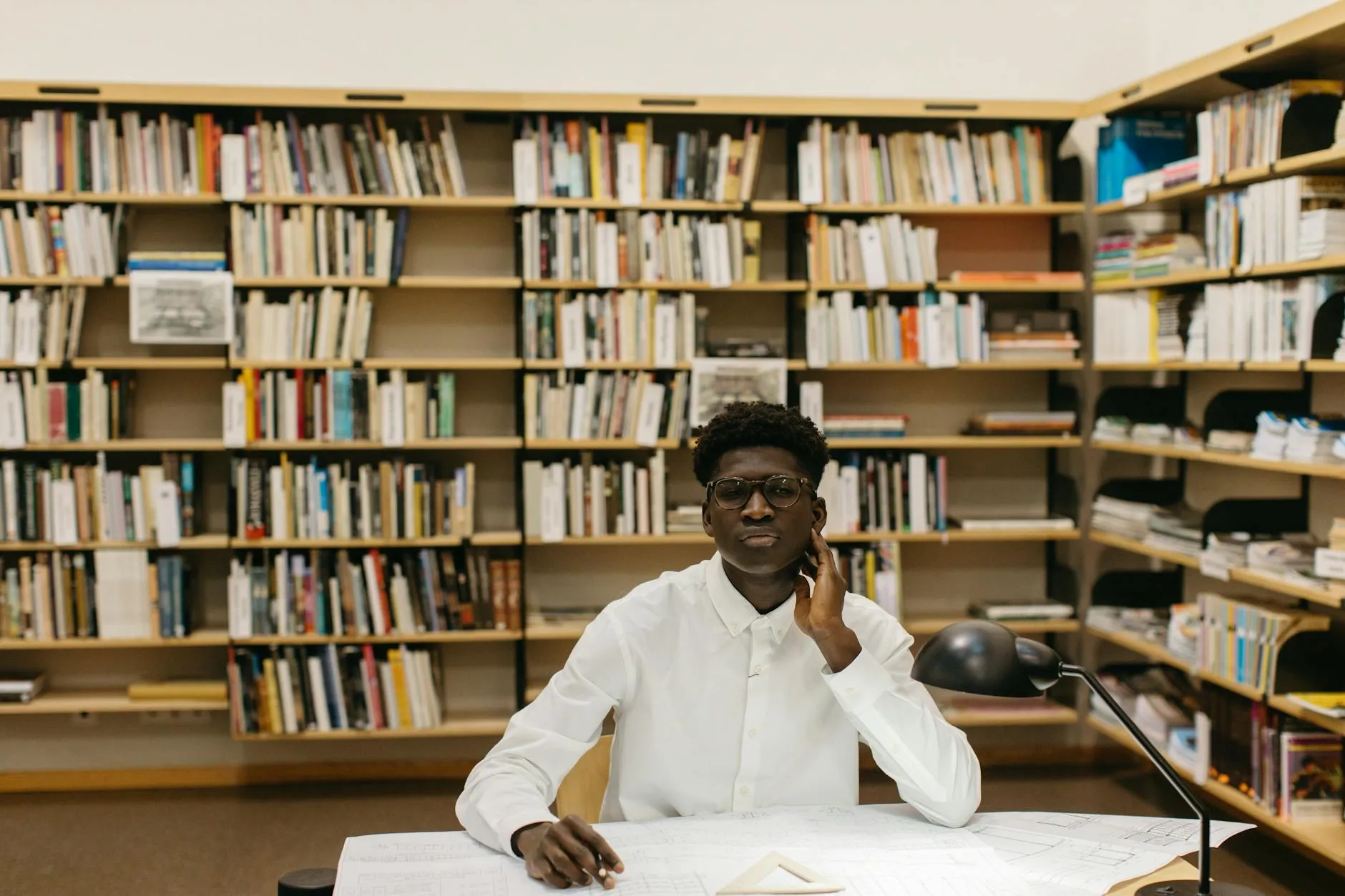 Estudante negro em biblioteca universitária cercado de livros, olhando pensativo