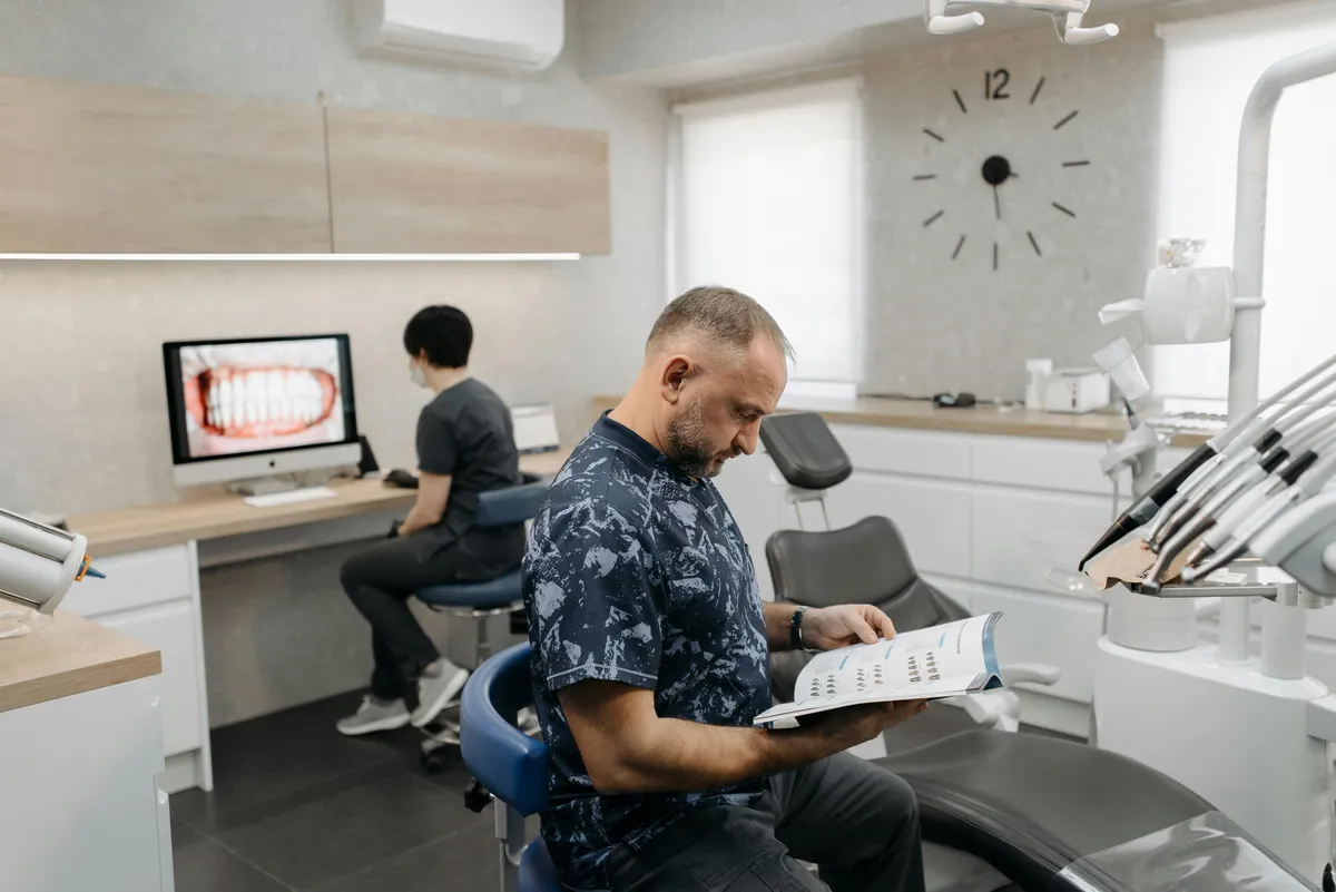 Profissional de odontologia lendo um folheto enquanto colega trabalha no computador em um consultório